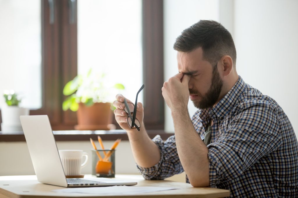 This is a picture of a man at desk rubbing the bridge of his nose and eyes because of headaches. His neck pain is causing his headache.