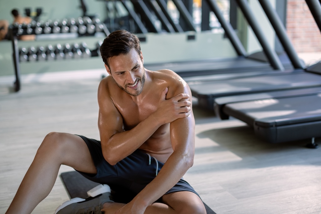 Person experiencing shoulder pain after working out at a gym in Edmonton. It depicts the title of the post 'Why Does My Shoulder Hurt After Working Out?'